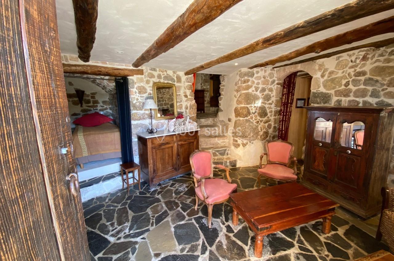 Salon rustique avec murs en pierre, poutres apparentes, mobilier en bois et fauteuils anciens, vue sur une chambre.