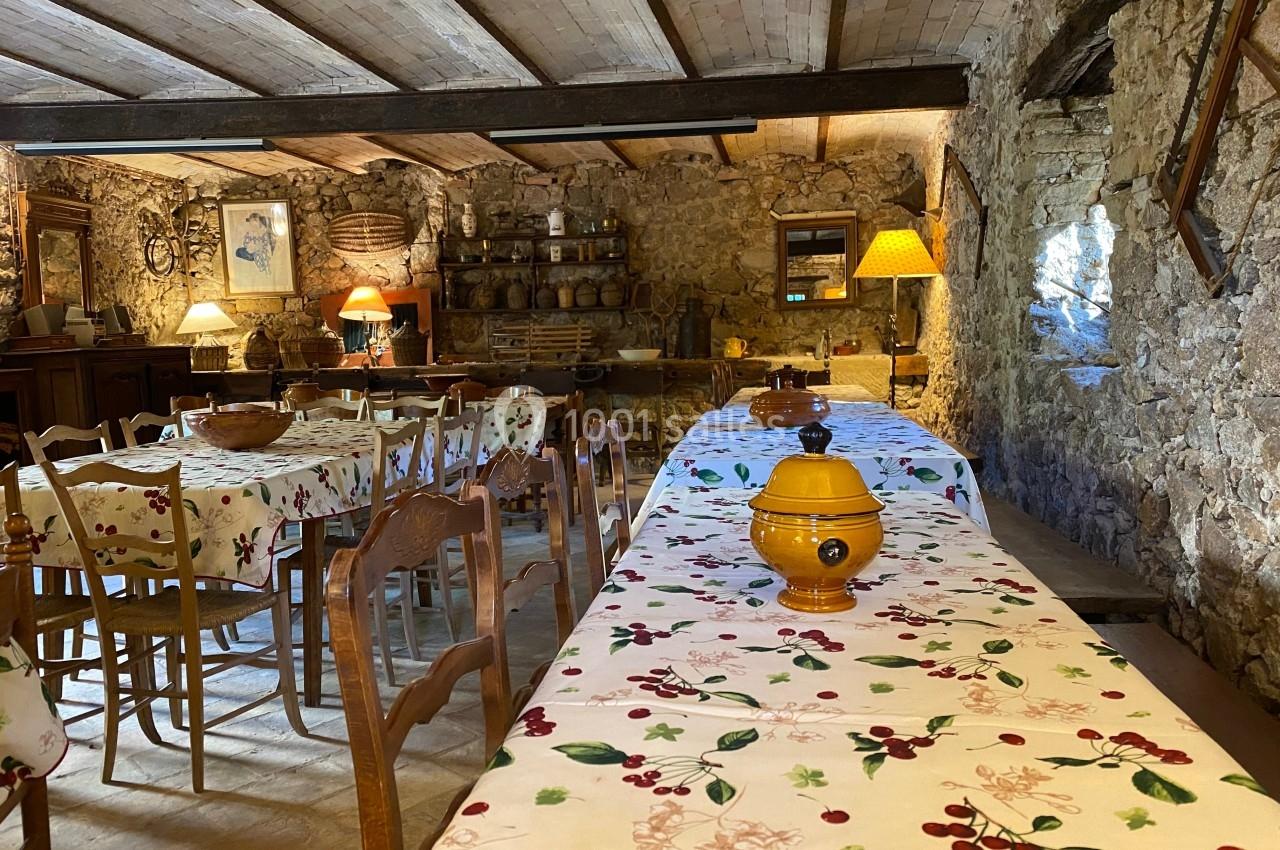Salle rustique avec tables couvertes de nappes à motifs de cerises, éclairée par des lampes et des murs en pierre apparente.