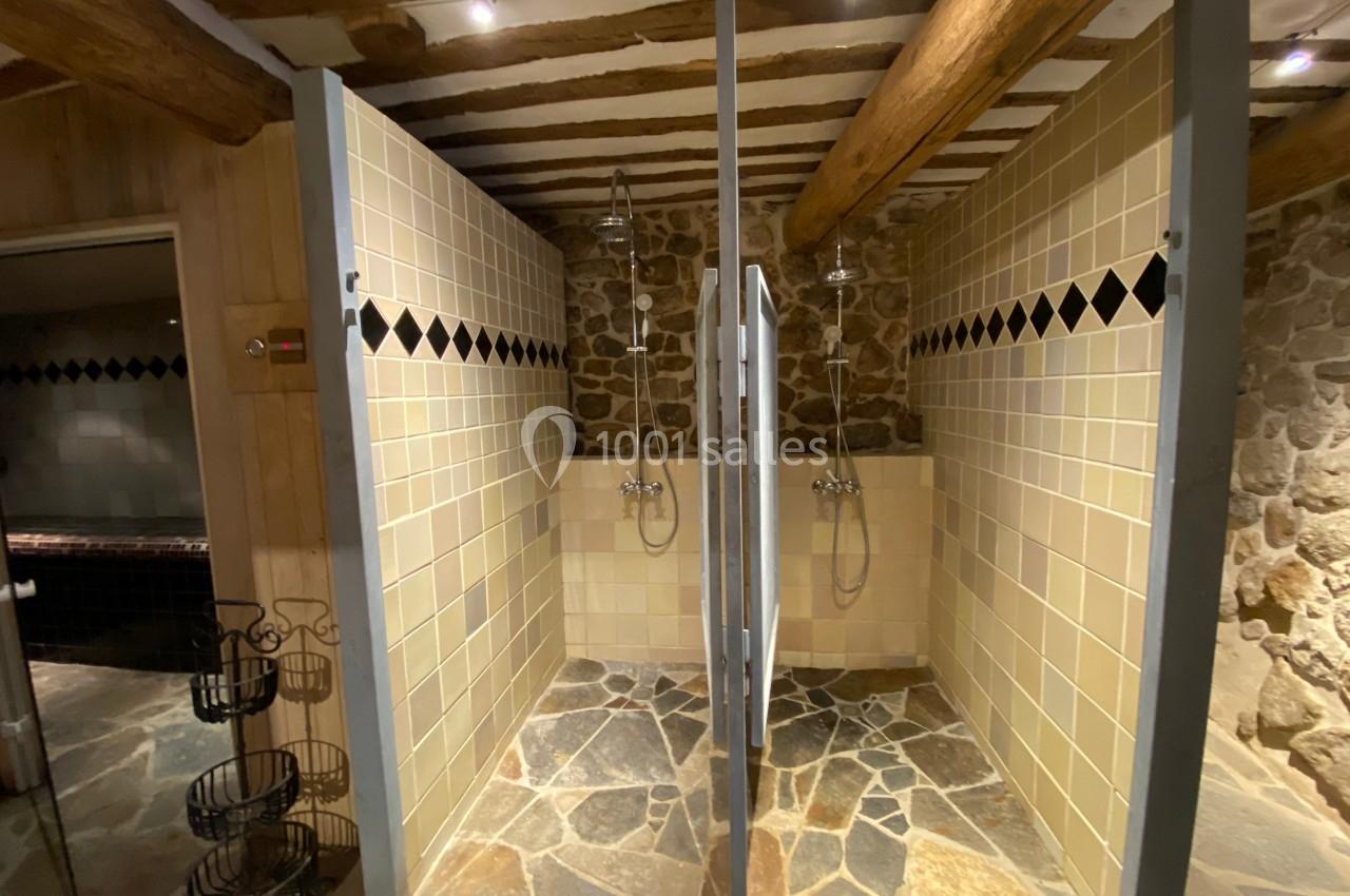 Douches en cabine séparées par des cloisons, avec murs en pierre et carrelage beige sous un plafond en poutres apparentes.