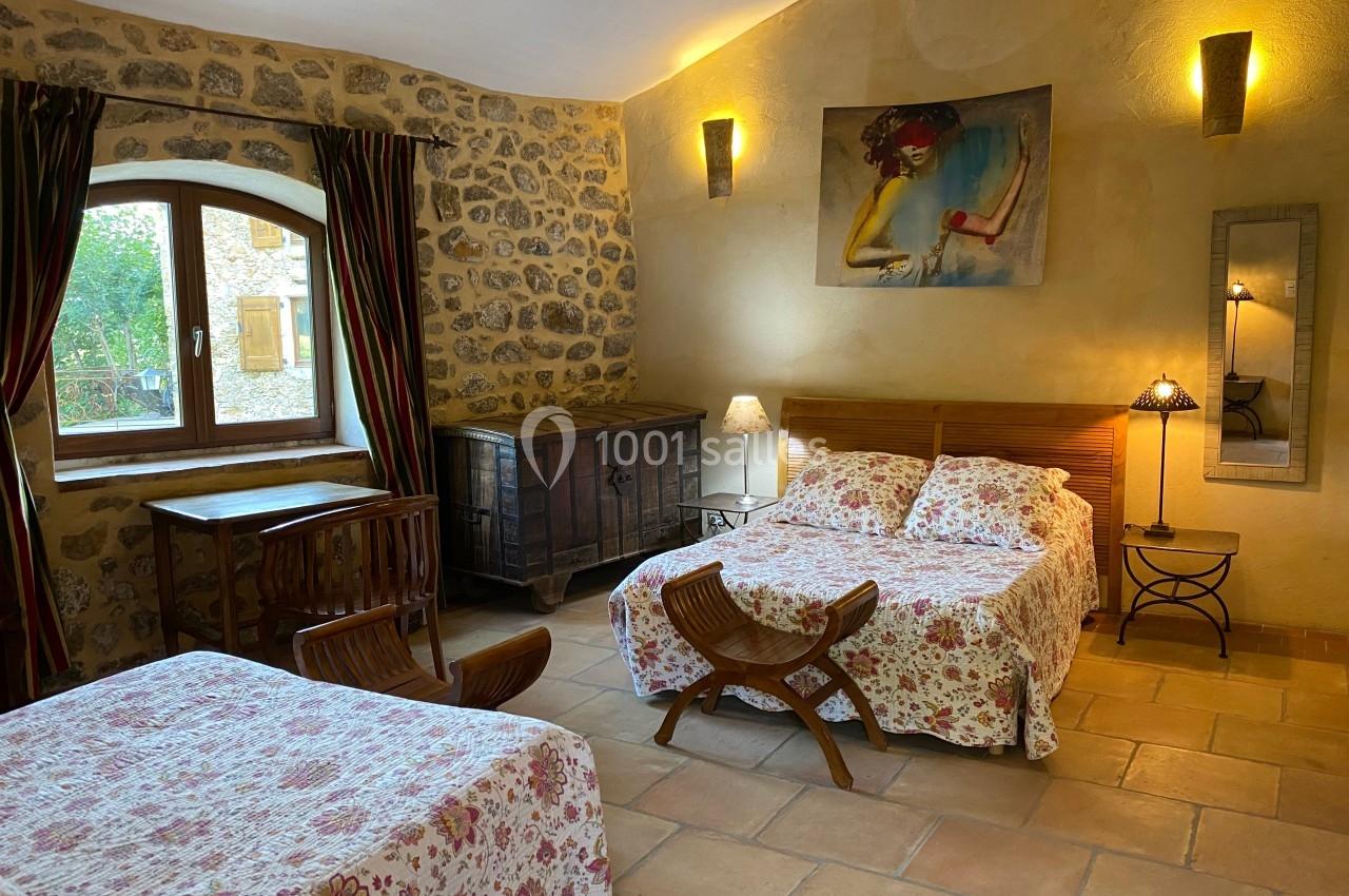 Chambre rustique avec murs en pierre, lits à motifs fleuris, mobilier en bois et lumière naturelle provenant d'une fenêtre.