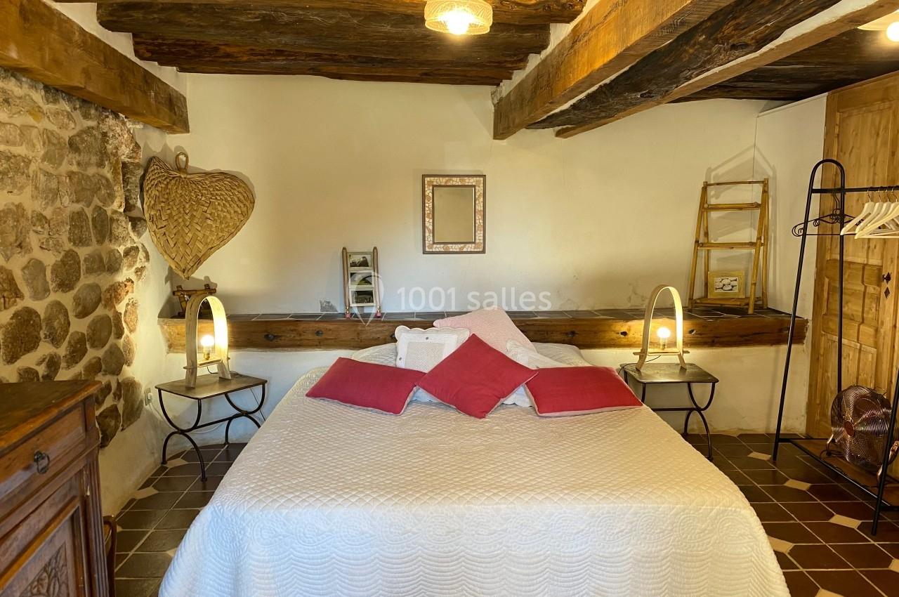 Chambre rustique avec poutres apparentes, lit double avec couvre-lit blanc et coussins roses, éclairée par deux lampes.
