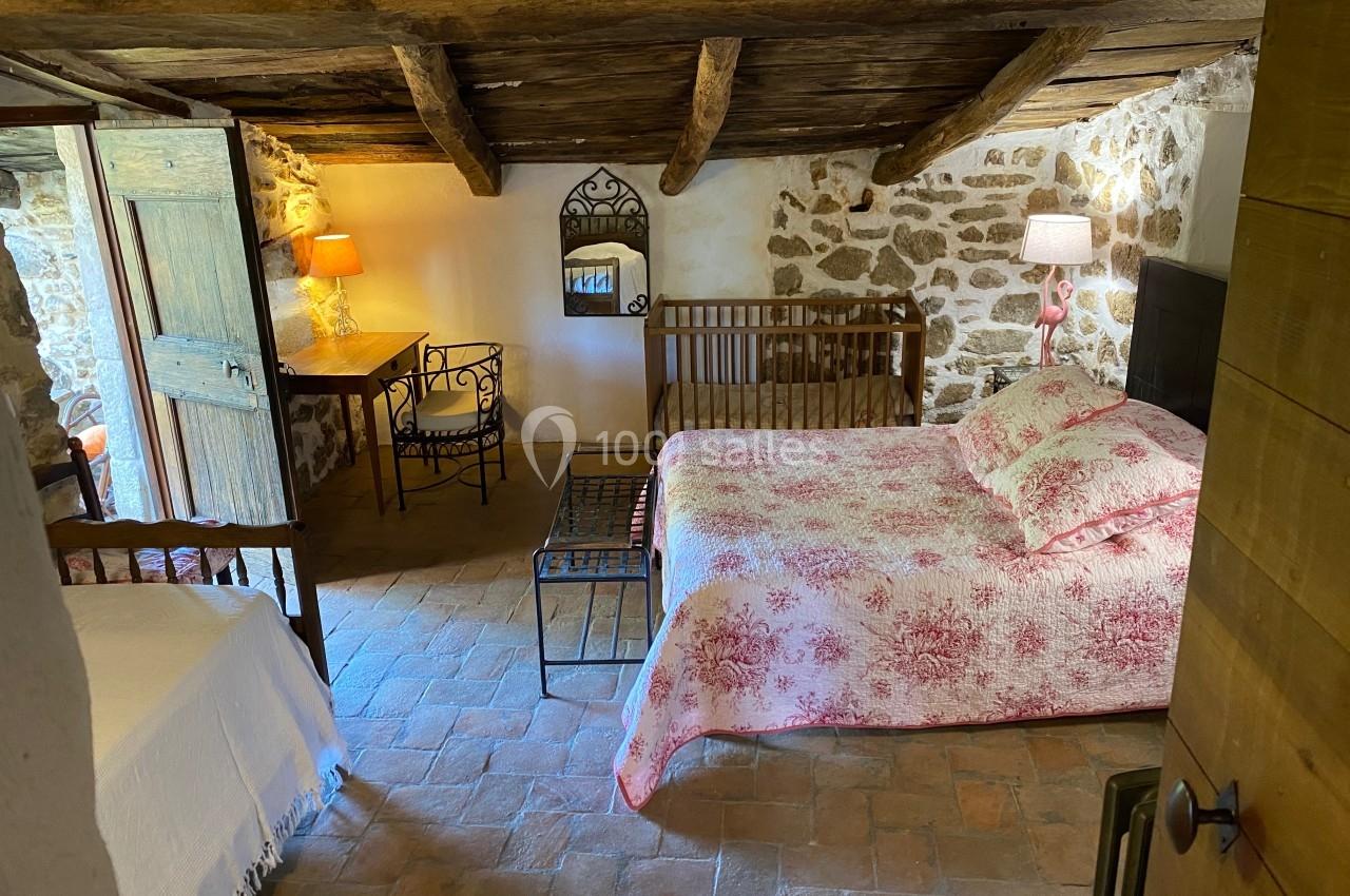 Chambre rustique avec murs en pierre, poutres apparentes, lit double, lit simple, bureau et éclairage chaleureux.