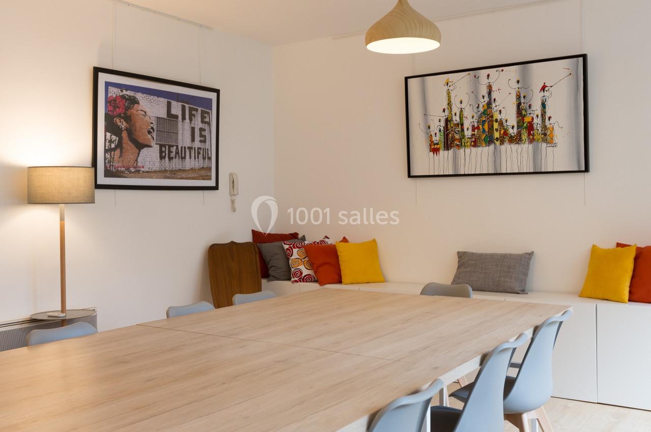 Salle à manger lumineuse avec une grande table en bois, chaises grises, coussins colorés et tableaux décoratifs au mur.