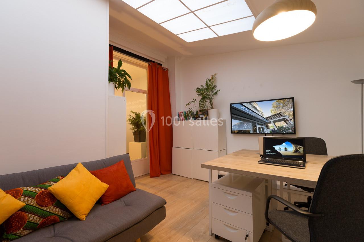 Bureau moderne avec canapé, table de travail, écran mural, plantes et lumière naturelle provenant d'une fenêtre.