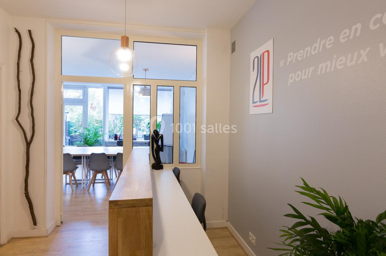 Vue d'un espace d'accueil moderne avec comptoir en bois, plantes et salle de réunion lumineuse en arrière-plan.