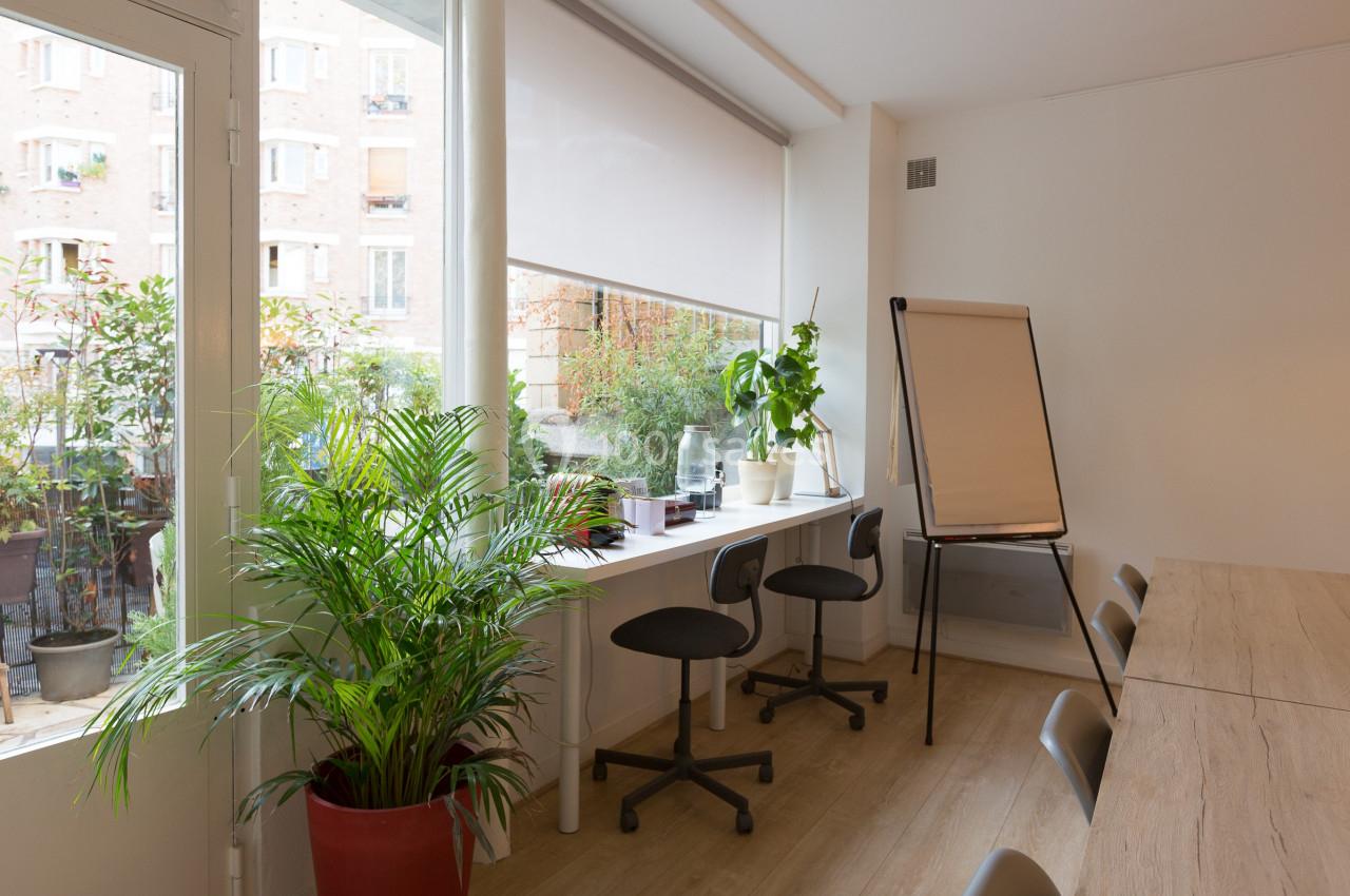 Salle lumineuse avec grandes fenêtres, plantes, bureau équipé de chaises et tableau de conférence sur pied.