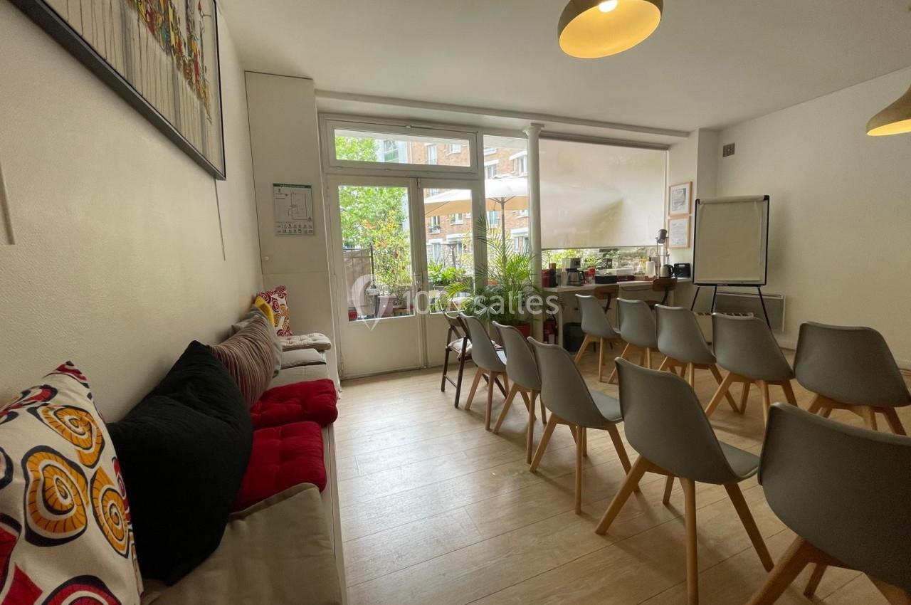Salle lumineuse avec chaises alignées, canapé coloré, tableau blanc et vue sur un jardin extérieur.