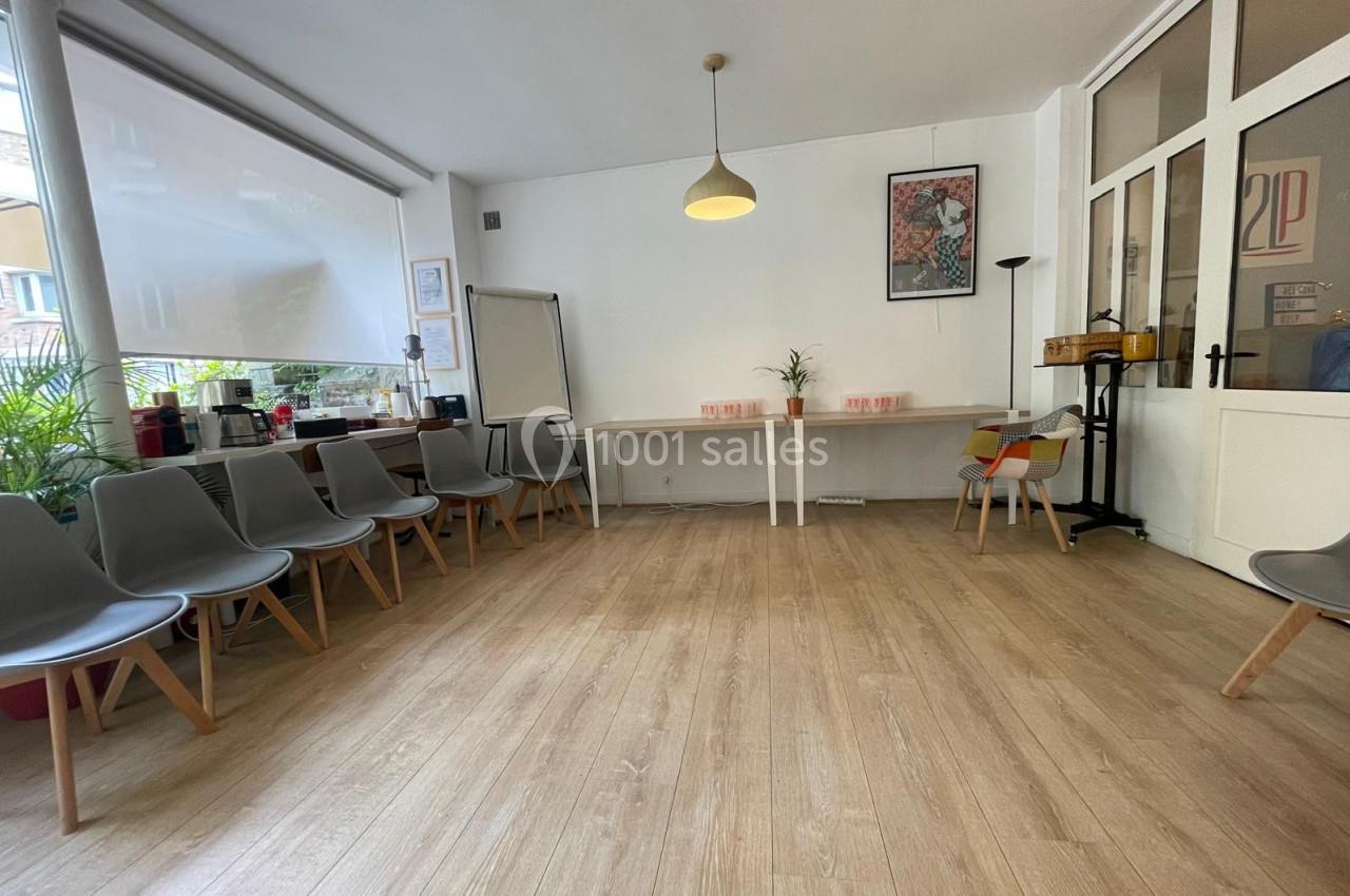 Salle lumineuse avec parquet, chaises disposées en cercle, tableau blanc, table et plantes en arrière-plan.