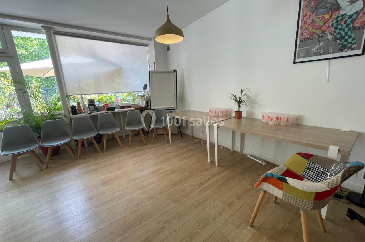 Salle lumineuse avec chaises alignées, tables en bois, plantes et un tableau de conférence près d'une grande fenêtre.