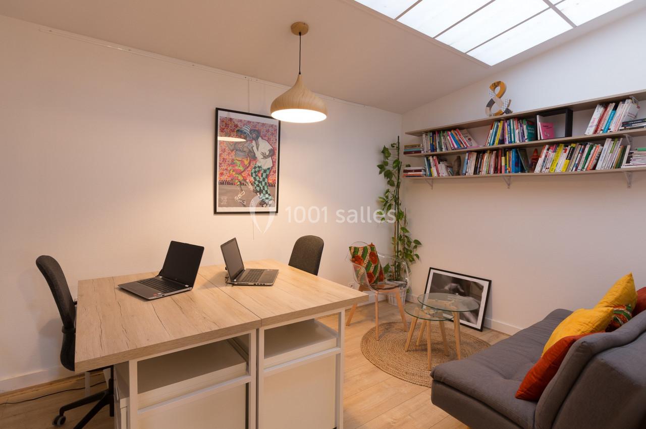 Bureau lumineux avec deux postes de travail, étagères remplies de livres, plantes et canapé dans un coin détente.