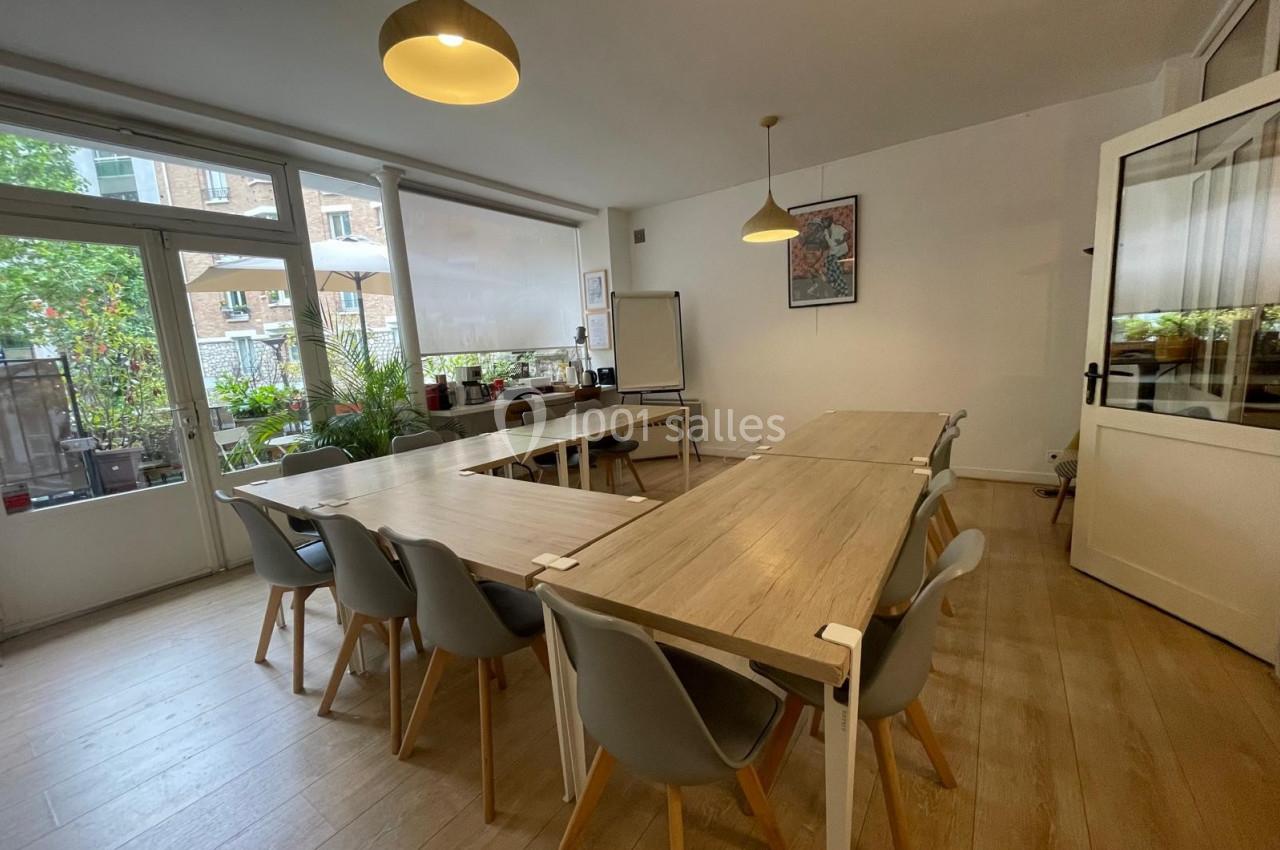 Salle lumineuse avec tables en bois, chaises grises et vue sur une terrasse arborée à travers de grandes baies vitrées.