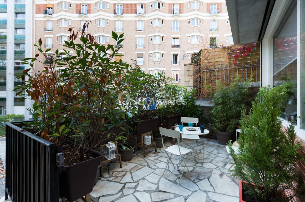 Terrasse en pierre avec table et chaises blanches, entourée de plantes en pots, donnant sur des immeubles en briques.
