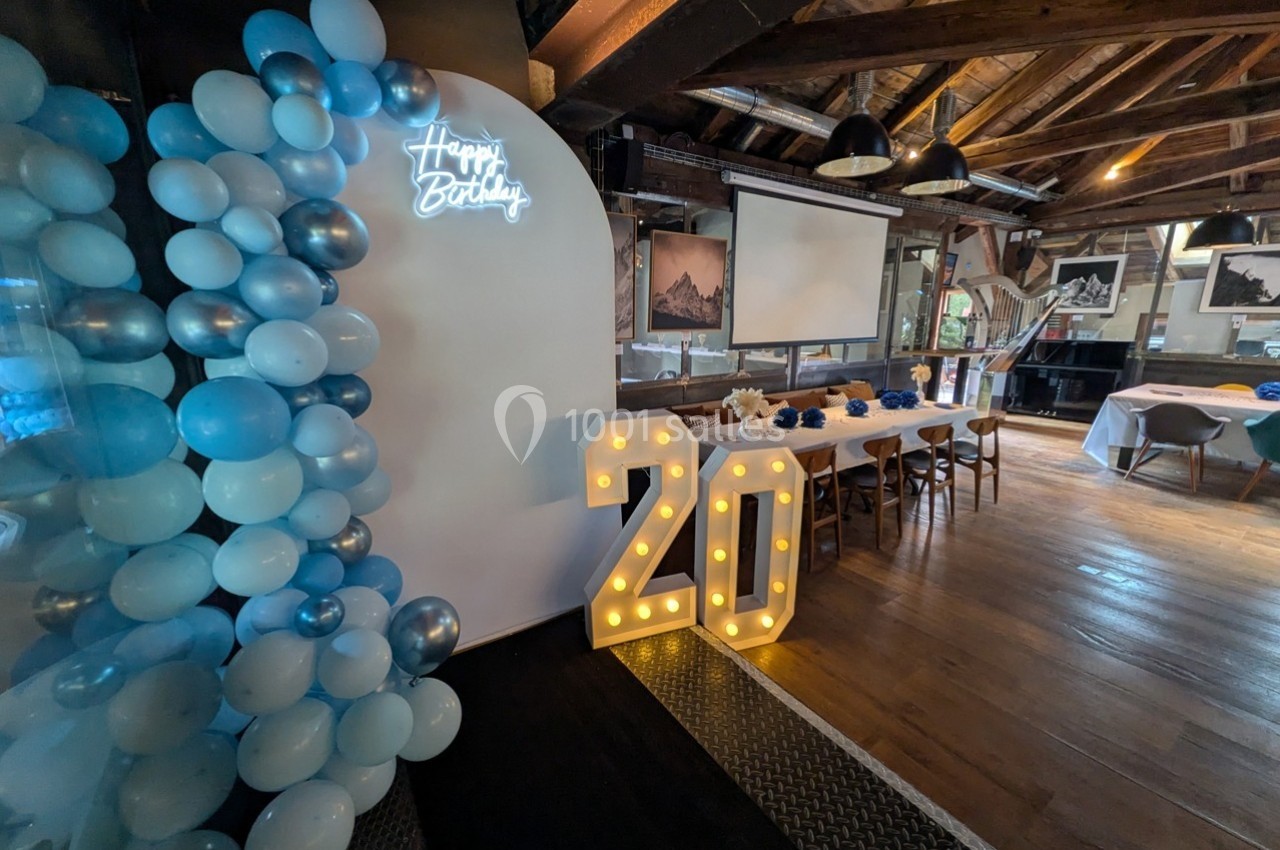 Décoration d'anniversaire avec ballons bleus, chiffre lumineux ’20’ et table dressée dans une salle en bois.