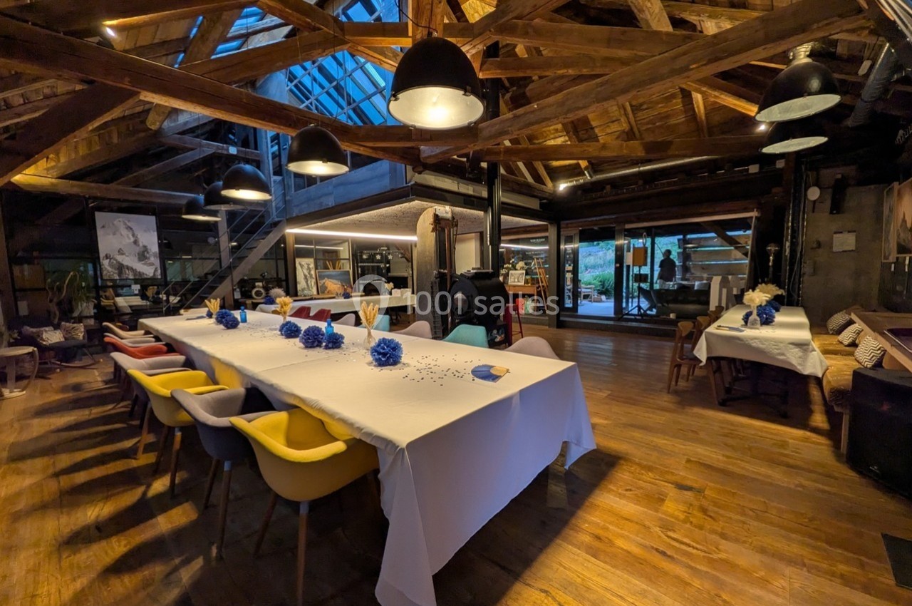 Grande salle en bois avec une longue table dressée, des chaises colorées et une lumière tamisée sous une charpente apparente.