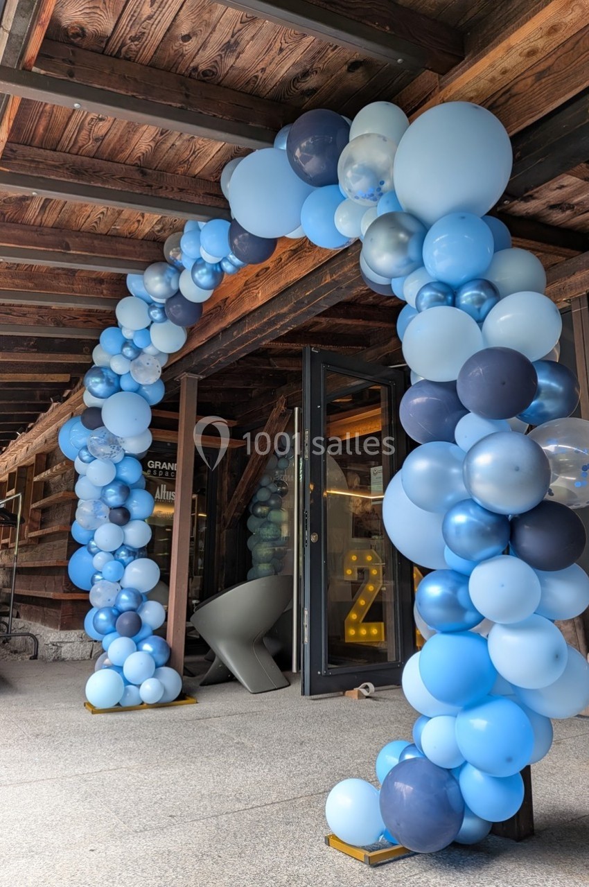 Arche décorative en ballons bleus et blancs installée à l'entrée d'un bâtiment en bois.