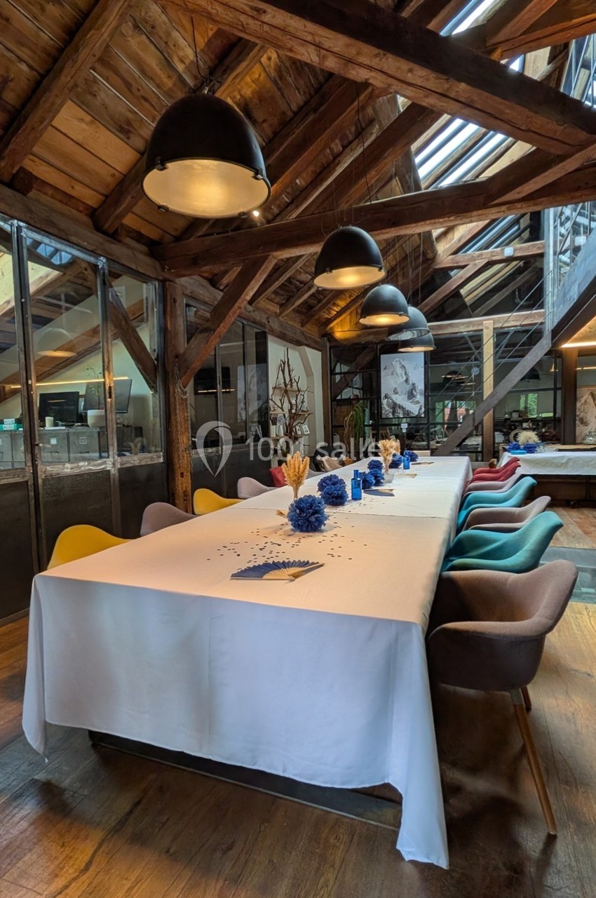 Grande table décorée de fleurs bleues dans une salle lumineuse avec charpente en bois et chaises colorées.