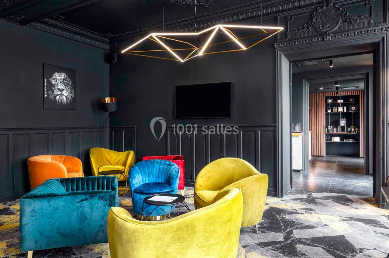 Salon moderne avec fauteuils colorés, murs noirs, lustre géométrique et sol en marbre, ouvert sur une autre pièce.