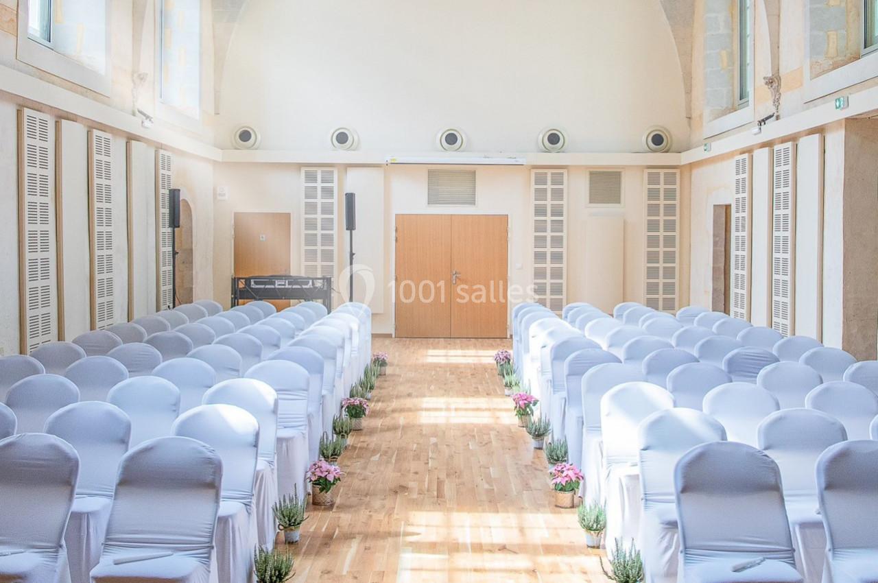 Salle lumineuse avec des rangées de chaises blanches alignées, décorée de plantes et fleurs le long de l'allée centrale.