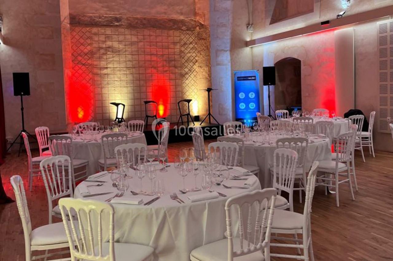 Salle de réception avec tables rondes dressées, chaises blanches et éclairage coloré devant une petite scène.