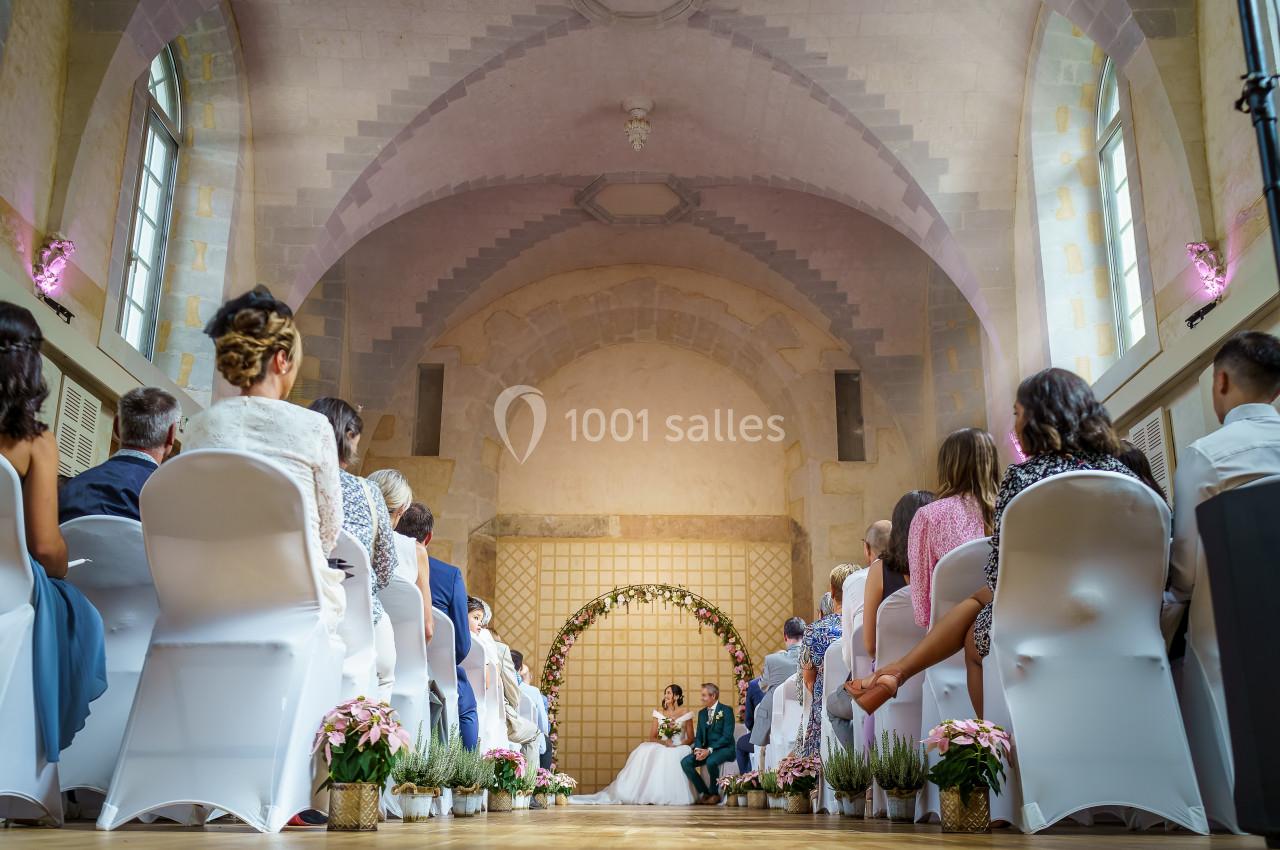 Un couple se marie sous une arche fleurie dans une salle voûtée, entouré d'invités assis.