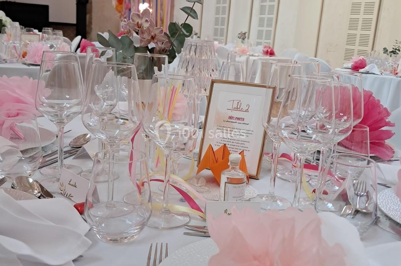 Table décorée pour un mariage avec fleurs roses, verres, menus et guirlandes lumineuses en arrière-plan.