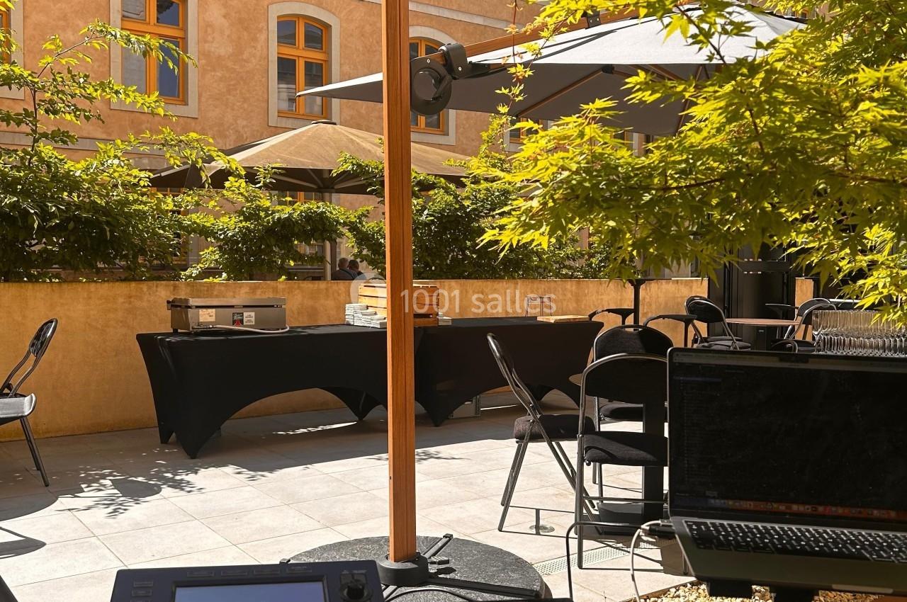 Console DJ en premier plan sur une terrasse ensoleillée, avec tables, parasols et bâtiment en arrière-plan.