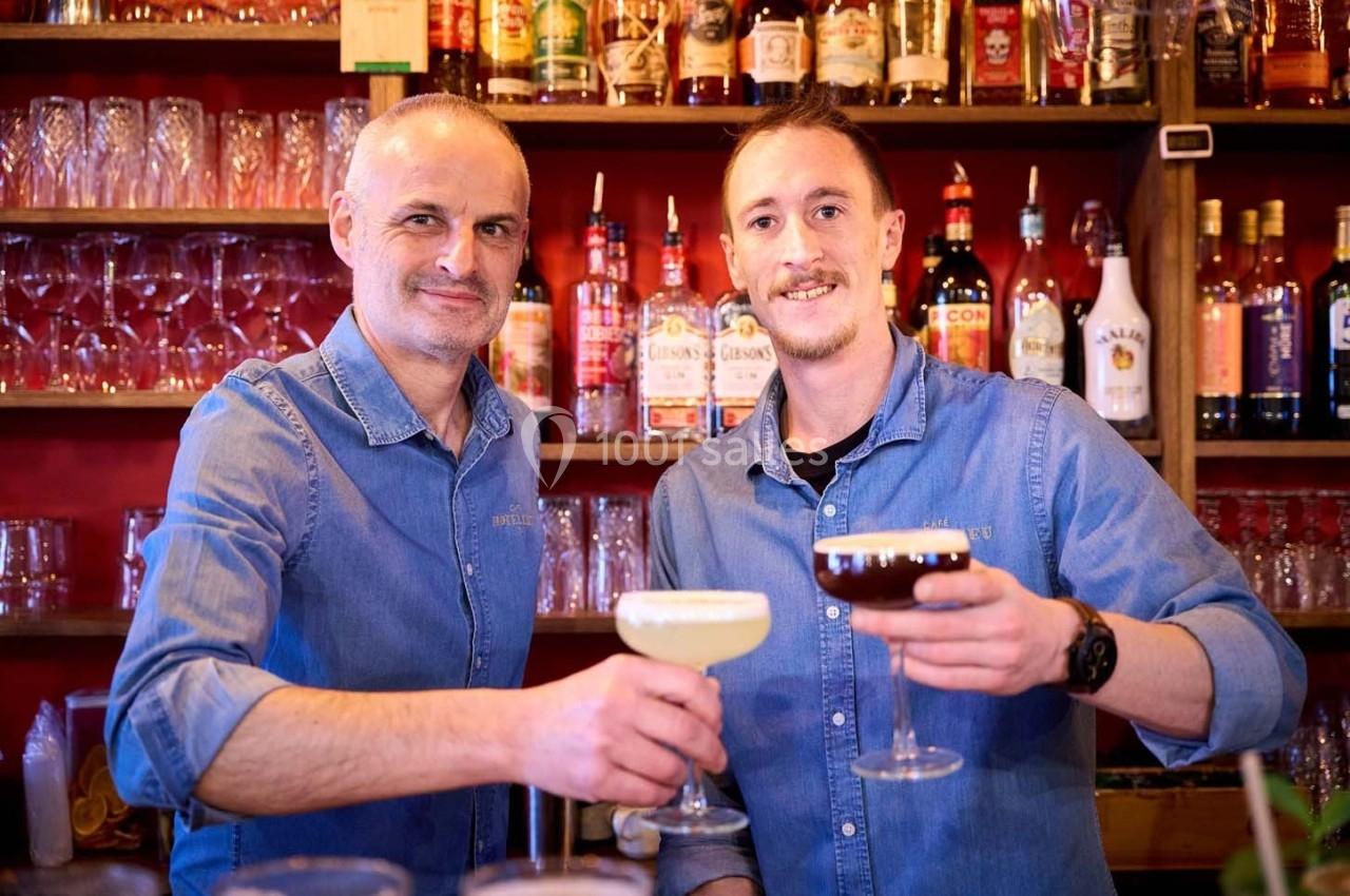 Deux hommes souriants en chemises en jean trinquent avec des cocktails dans un bar coloré.