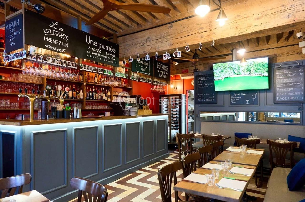 Intérieur d'un restaurant avec bar en bois, étagères de bouteilles, tables dressées et écran affichant un paysage.