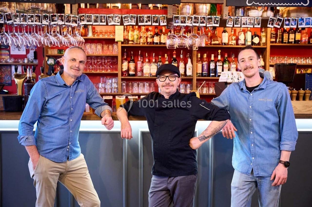 Trois hommes posent devant un bar avec des étagères remplies de bouteilles et des verres suspendus au-dessus du comptoir.