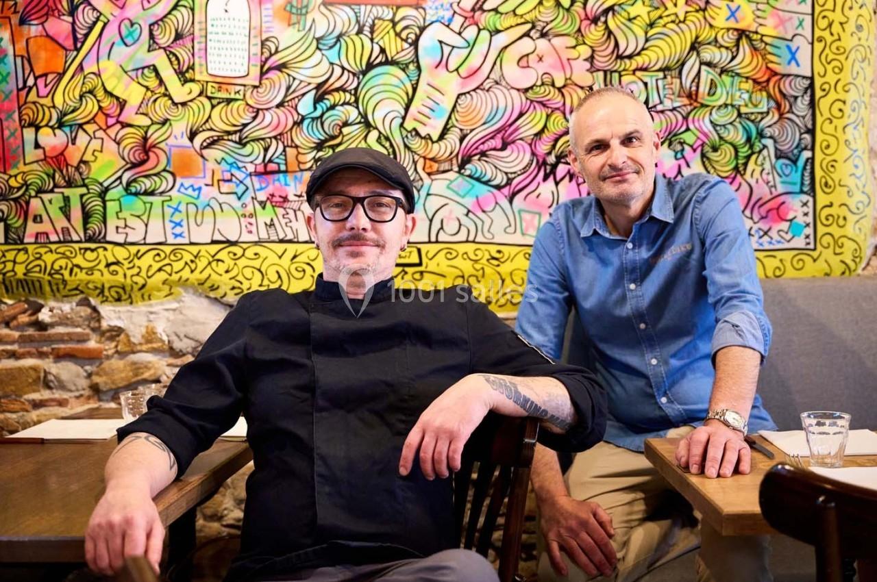 Deux hommes assis dans un café devant un mur coloré décoré de graffitis artistiques.