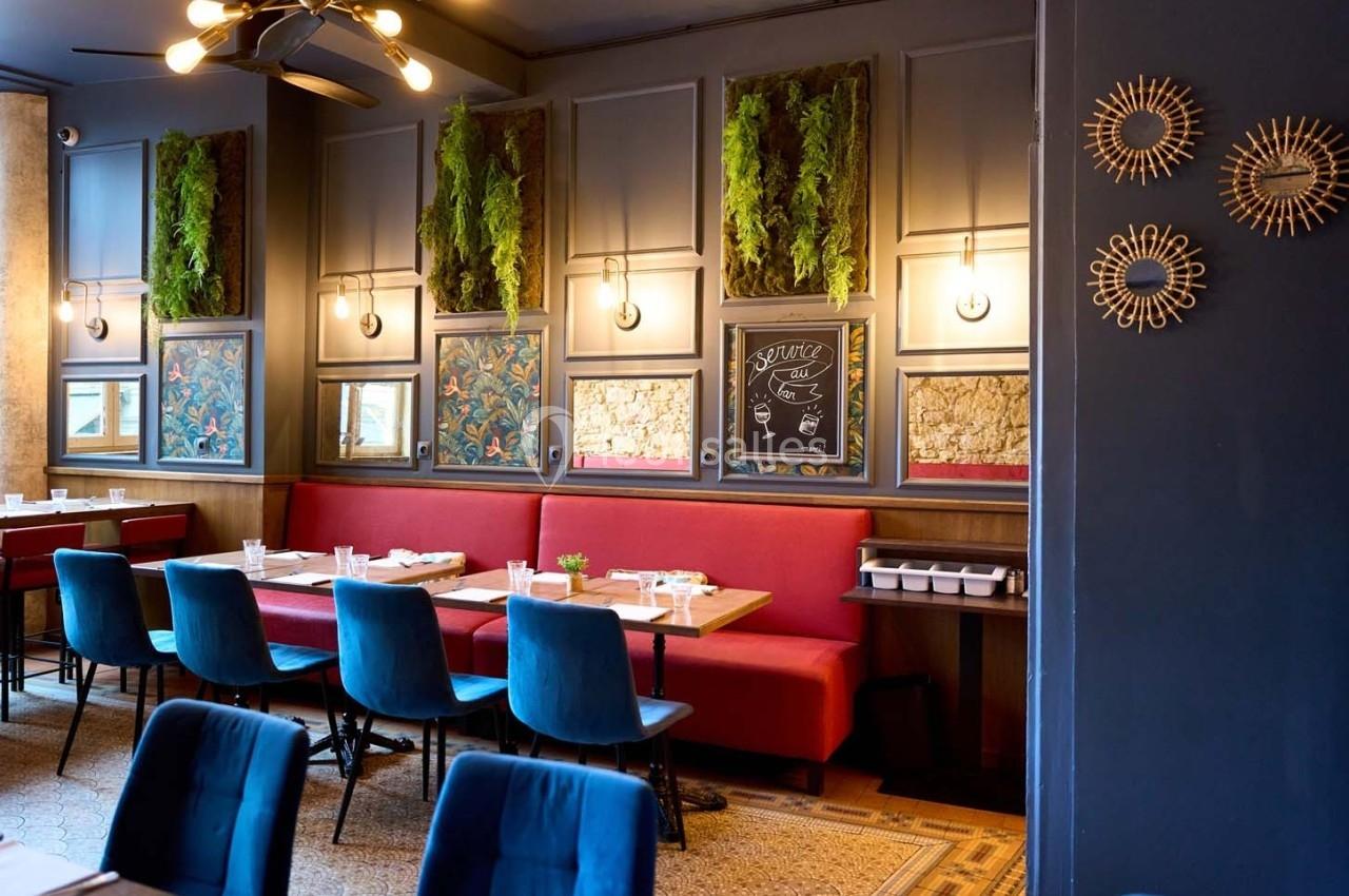 Salle de restaurant avec banquettes rouges, chaises bleues, décor végétal mural et éclairage chaleureux.