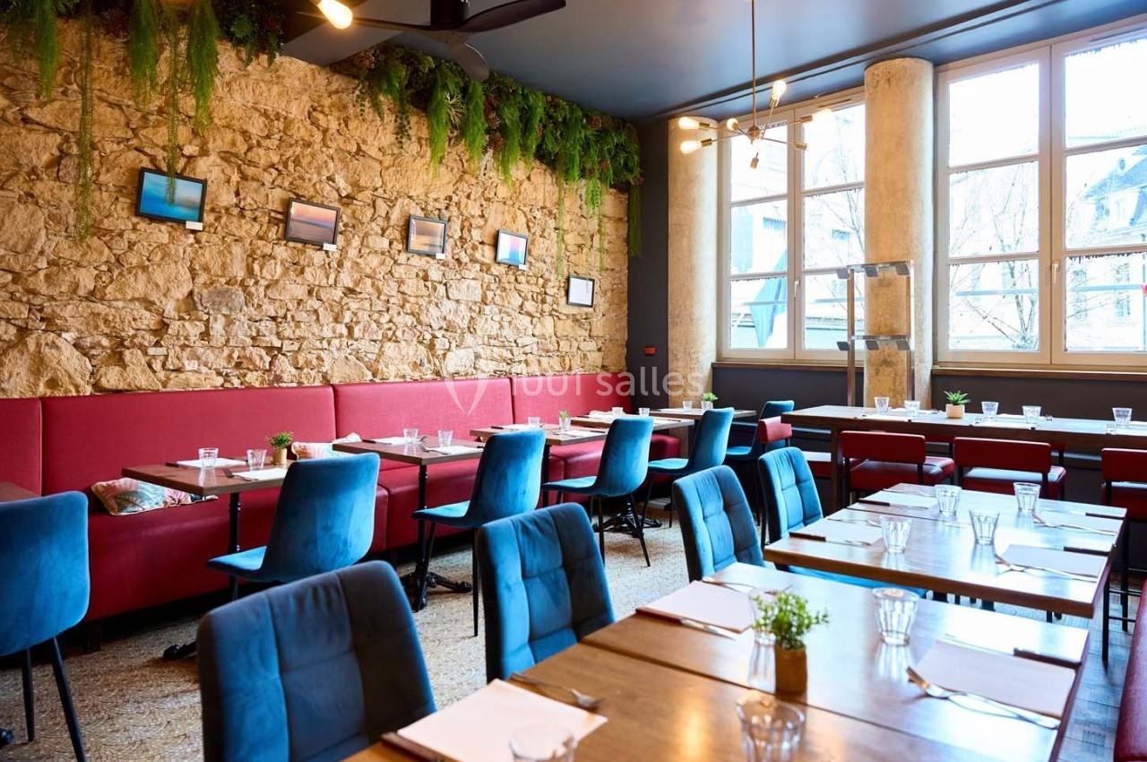Salle de restaurant lumineuse avec murs en pierre, banquettes rouges, chaises bleues et tables dressées.