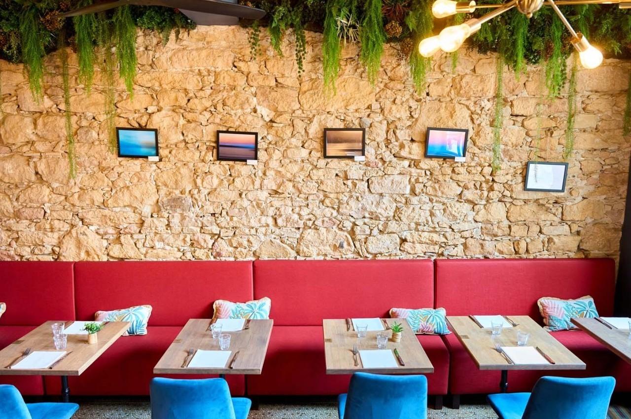 Mur en pierre décoré de cadres, banquette rouge, tables en bois et chaises bleues dans un espace lumineux.