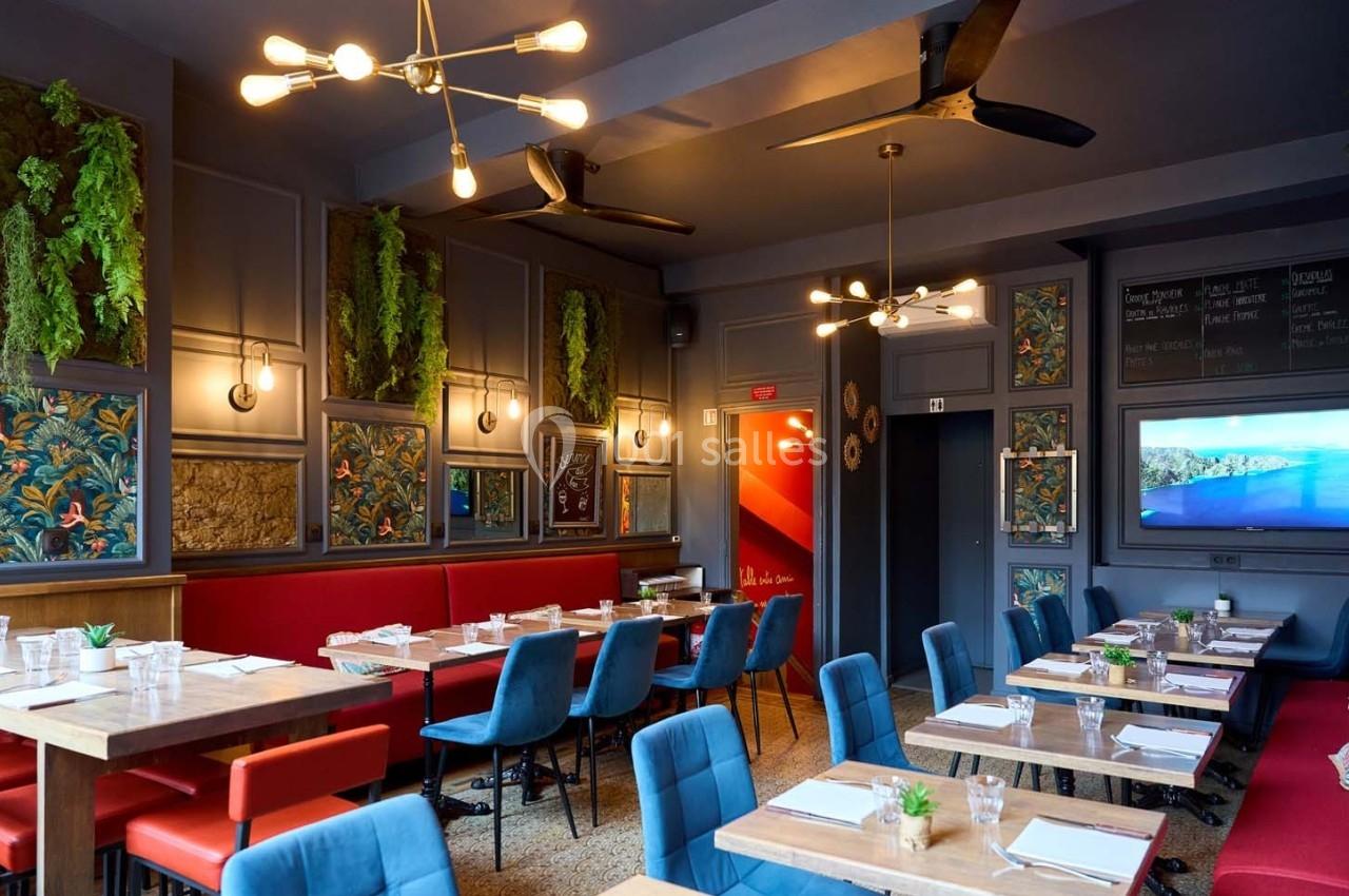 Salle de restaurant moderne avec tables en bois, chaises bleues, banquettes rouges et décor végétal sur les murs.
