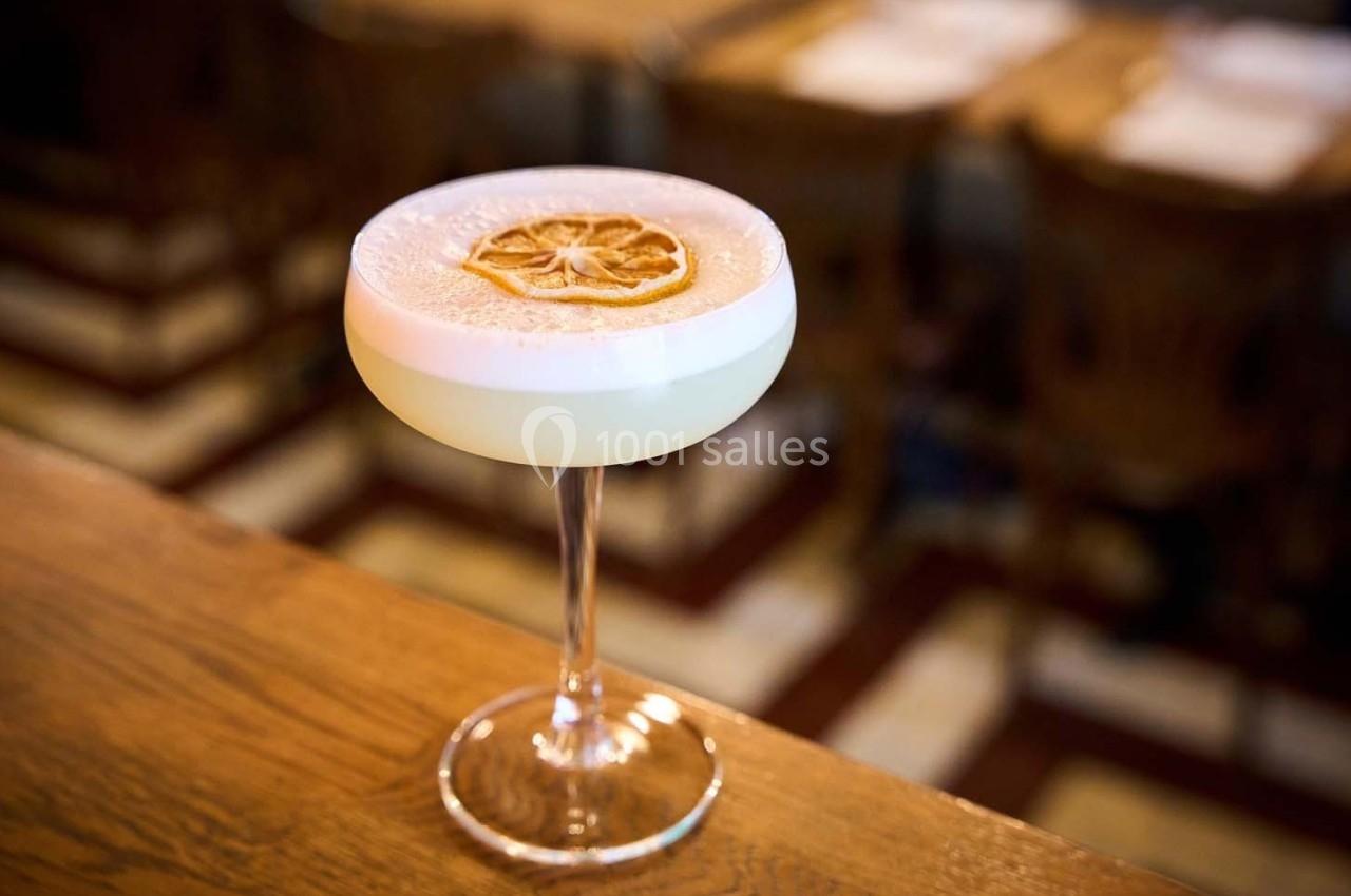 Verre à cocktail rempli d'une boisson mousseuse, garni d'une tranche d'agrume séchée, posé sur un comptoir en bois.