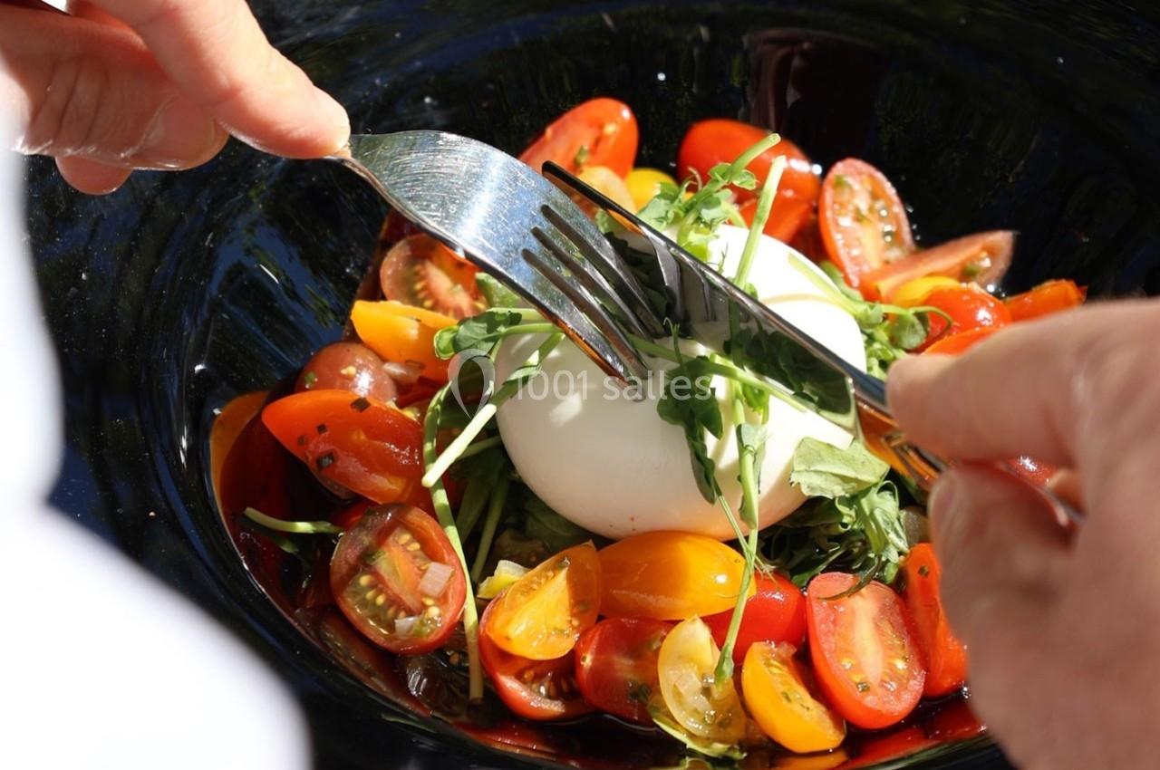 Personne coupant une burrata accompagnée de tomates cerises colorées et de jeunes pousses dans une assiette noire.