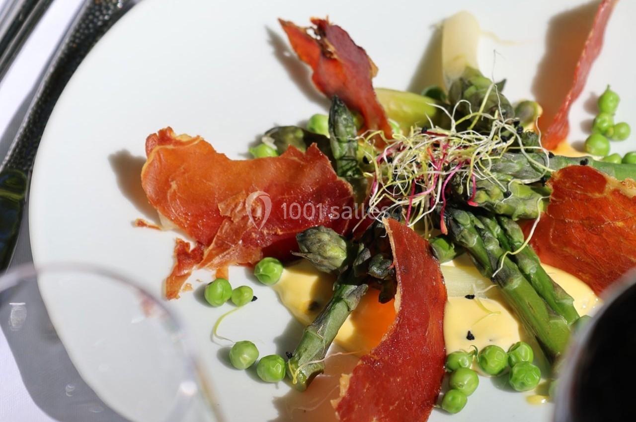 Assiette garnie d'asperges, petits pois, jambon croustillant, germes et sauce crémeuse, présentée sur une table.
