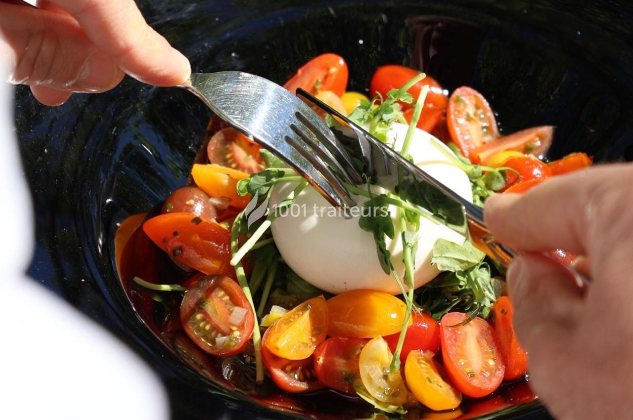 Personne coupant une burrata garnie de tomates cerises colorées et de jeunes pousses dans une assiette noire.