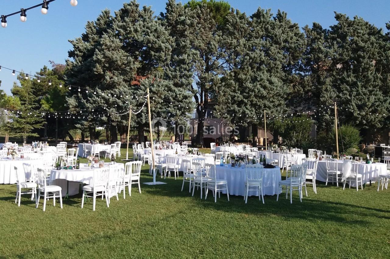 Tables et chaises blanches disposées en extérieur sur une pelouse, entourées d'arbres et décorées de guirlandes lumineuses.