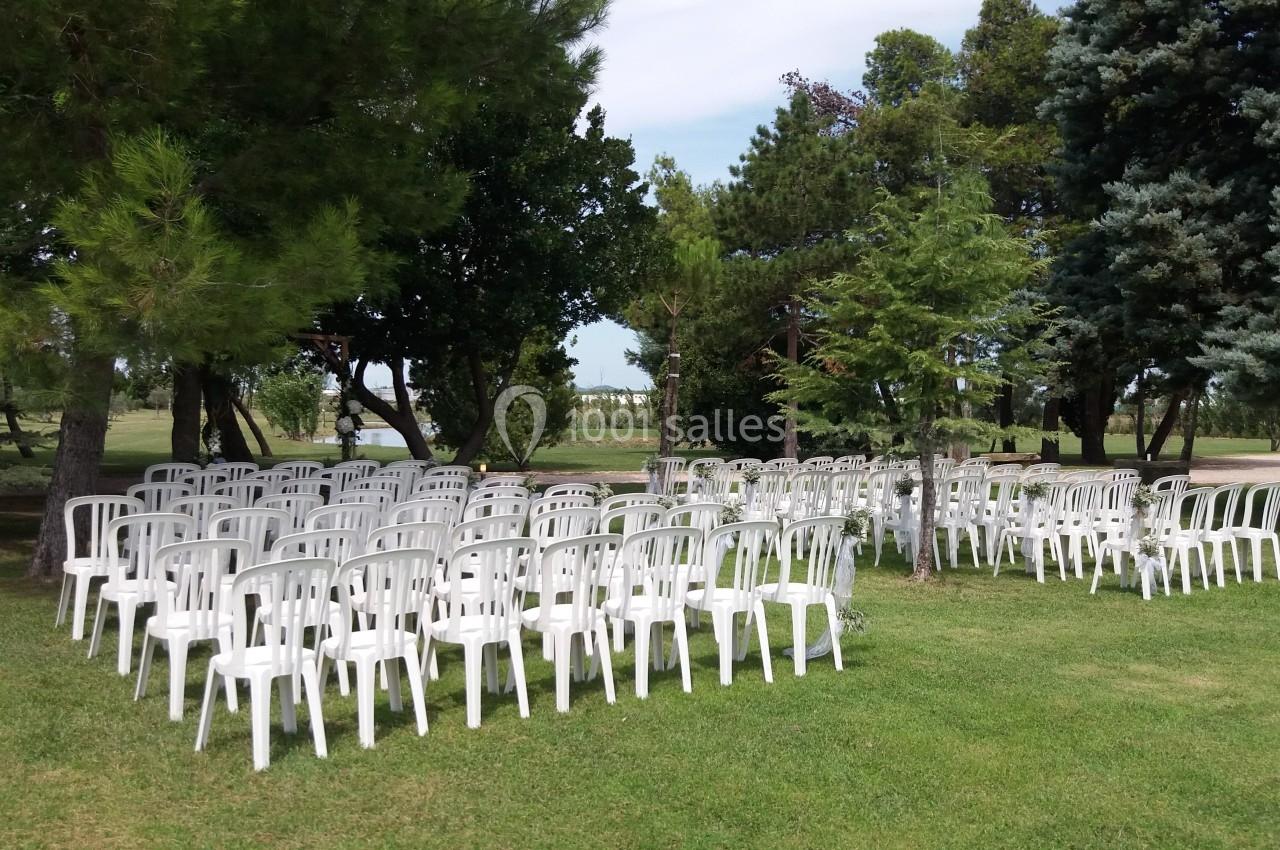 Chaises blanches en plastique disposées en rangées sur une pelouse, entourées d'arbres dans un espace extérieur.