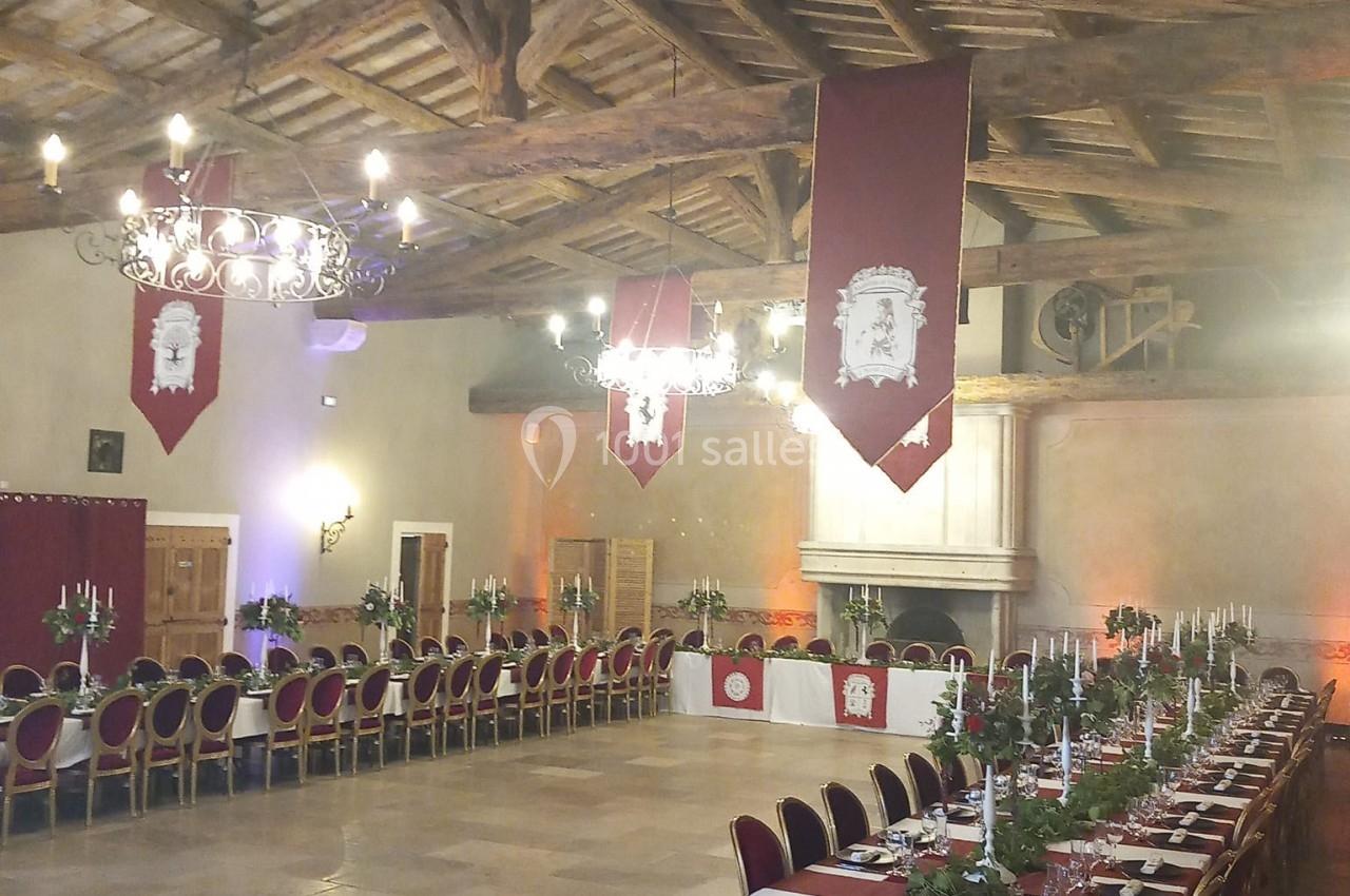 Grande salle de réception avec tables dressées, chandeliers, bannières rouges et plafond en bois apparent.