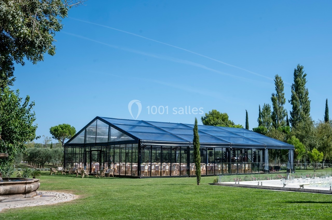 Structure en verre et métal dans un jardin verdoyant, entourée d'arbres, avec une fontaine et une piscine à proximité.
