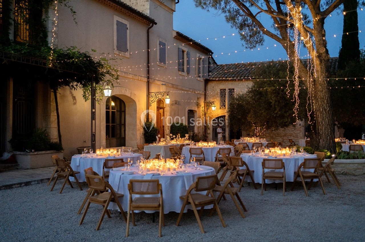 Tables rondes décorées pour un dîner en plein air, éclairées par des guirlandes lumineuses dans une cour au crépuscule.