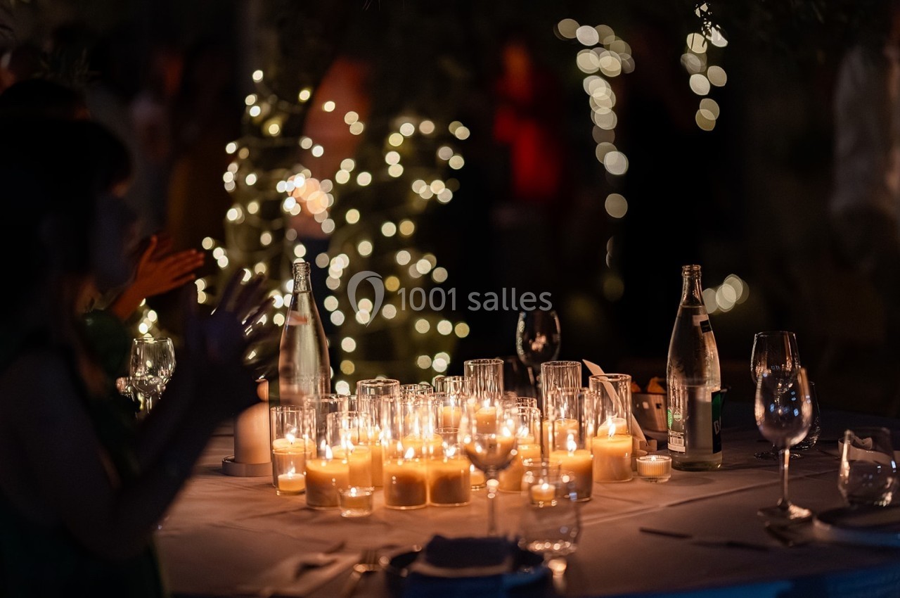 Bougies allumées sur une table de fête en soirée, entourées de verres et de bouteilles, avec des lumières floues en arrière…