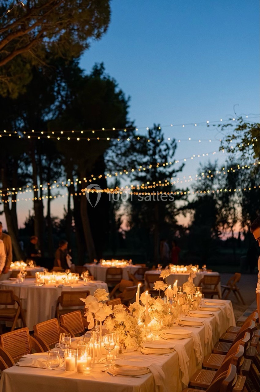 Tables décorées avec des bougies et des fleurs blanches sous des guirlandes lumineuses, en extérieur au crépuscule.
