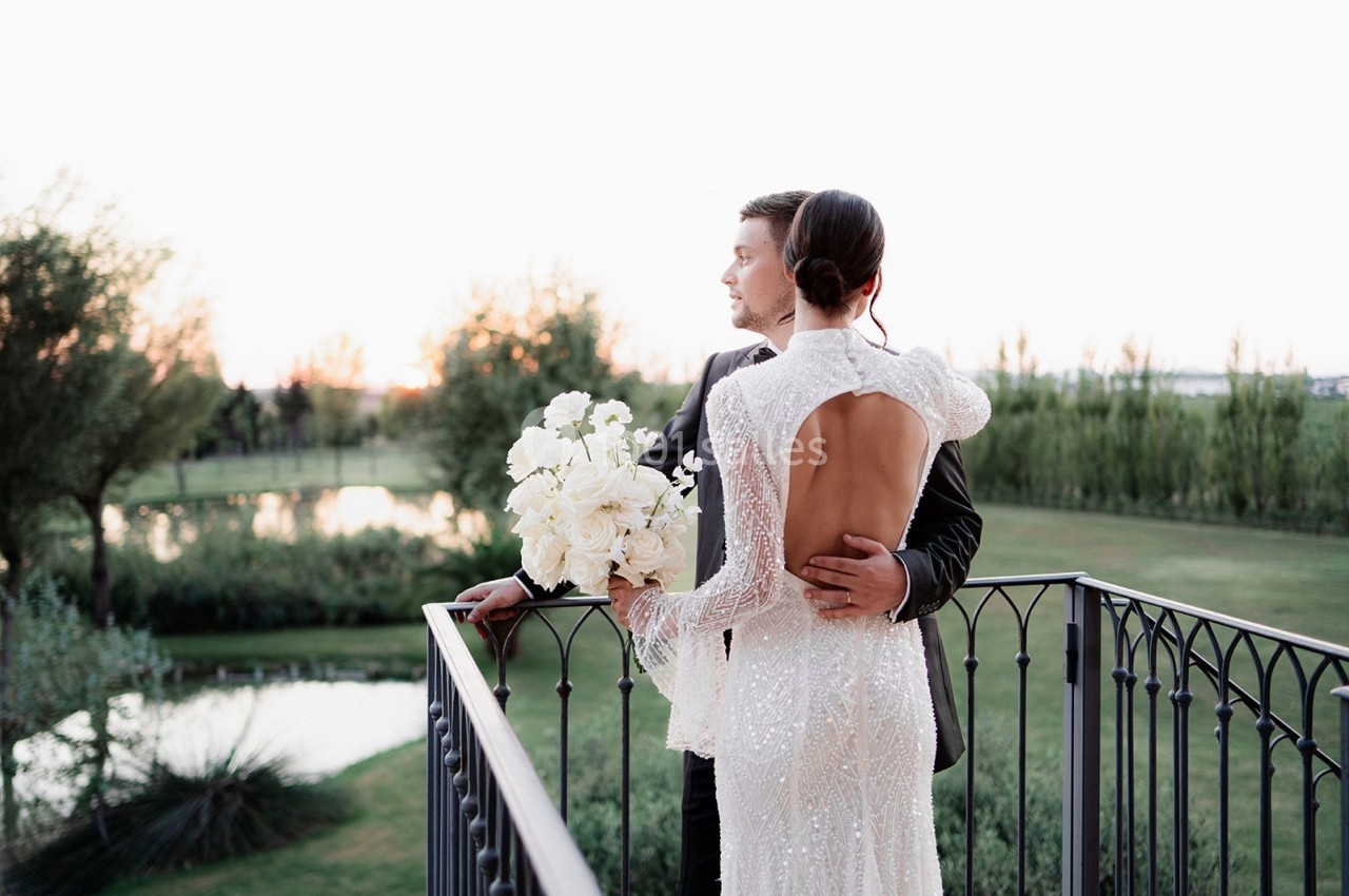 Un couple en tenue de mariage se tient sur un balcon, regardant un paysage verdoyant au coucher du soleil.