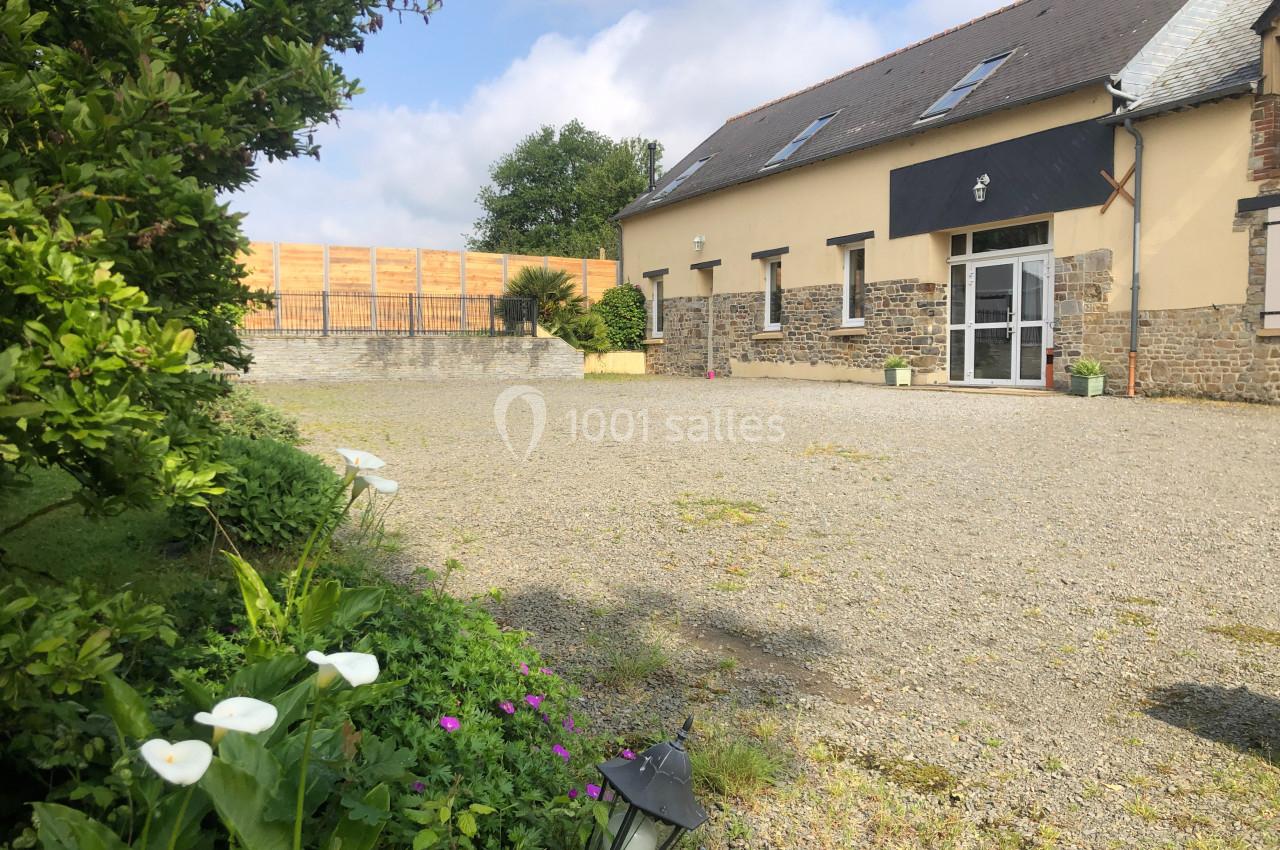 Cour gravillonnée devant un bâtiment en pierre avec des fleurs blanches et un arbuste au premier plan.
