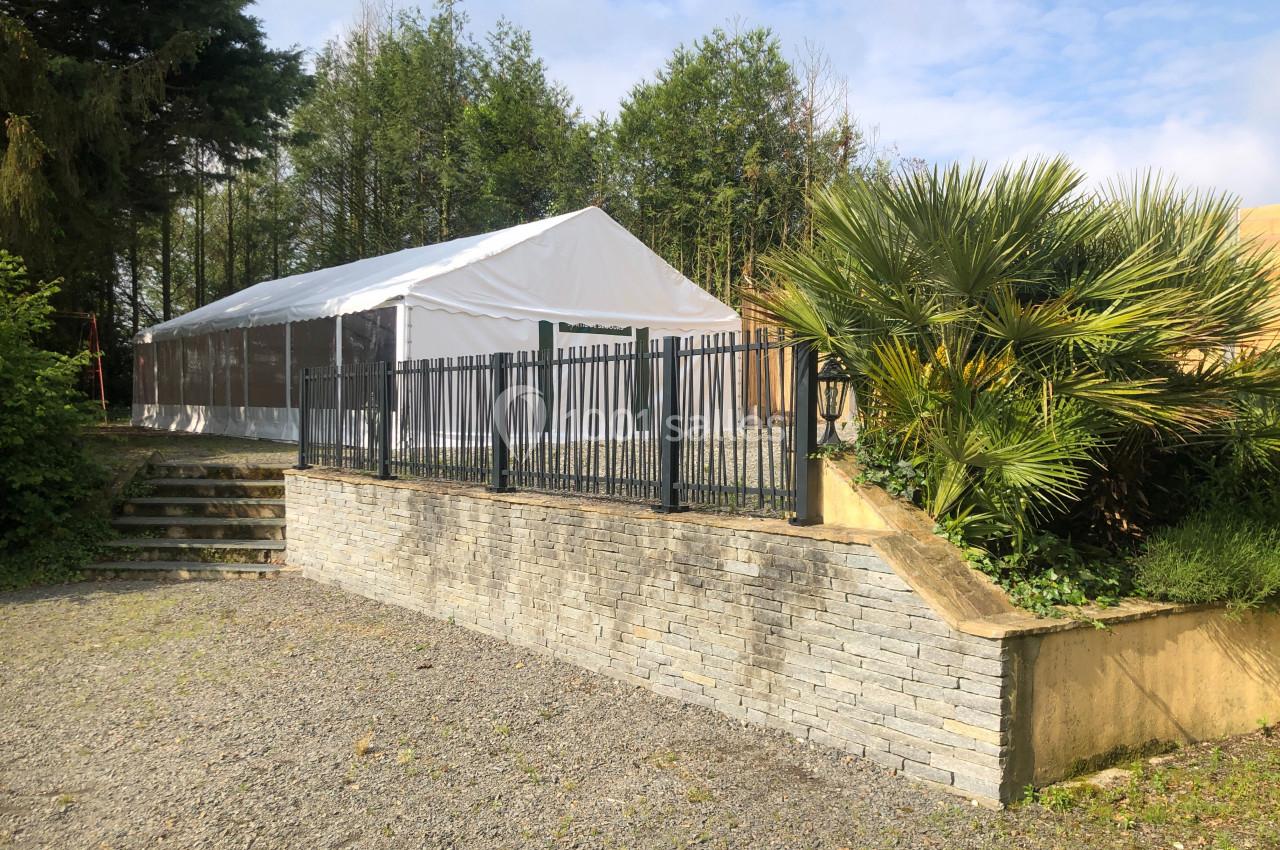 Tente blanche installée dans un jardin, entourée de végétation, avec une clôture en métal et un muret en pierre au premier…