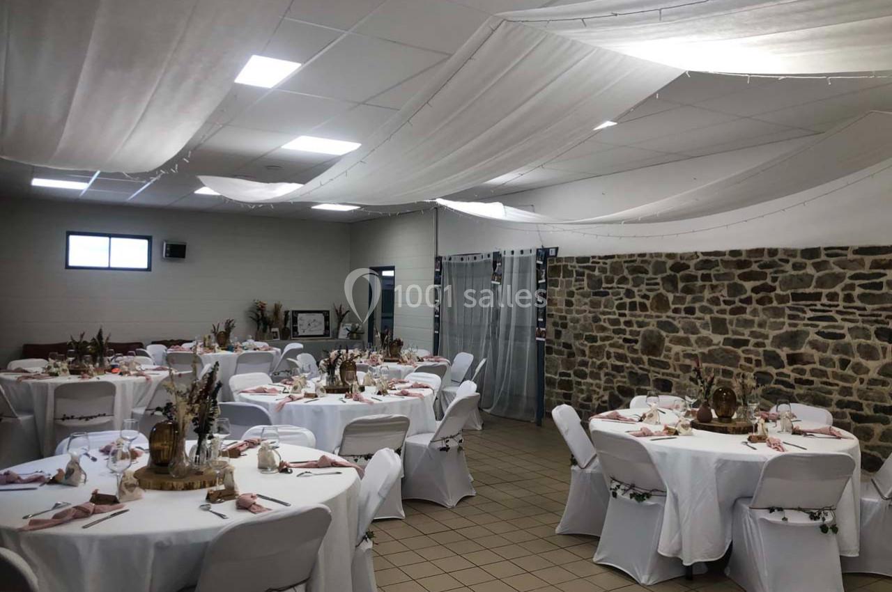 Salle décorée pour un événement, avec des tables rondes dressées, nappes blanches et décorations florales.