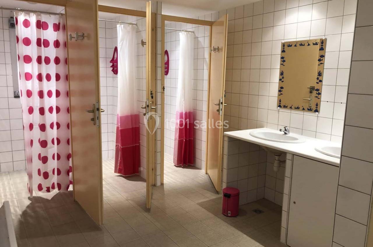 Vestiaire avec douches individuelles, rideaux à motifs roses et blancs, lavabo double et miroir mural.